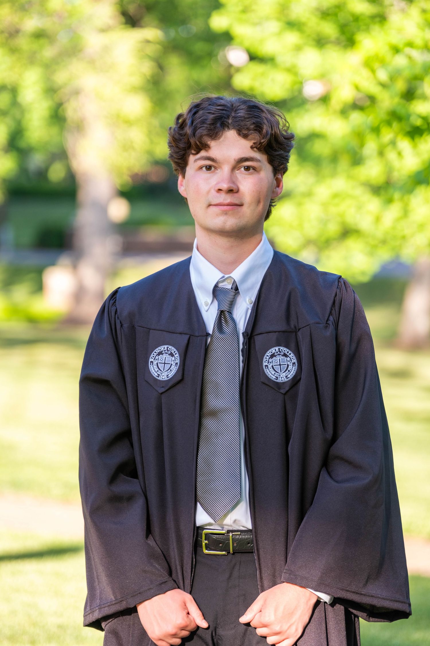 Young man in black graduation gown and gray tie posing outdoors with green trees in background.