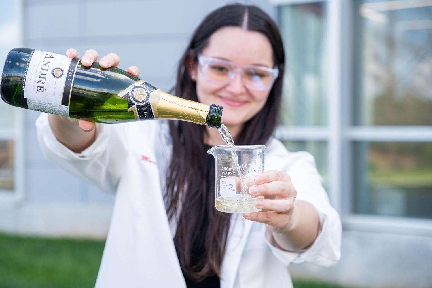 Female student in white lab coat pouring champagne into a glass beaker outdoors.
