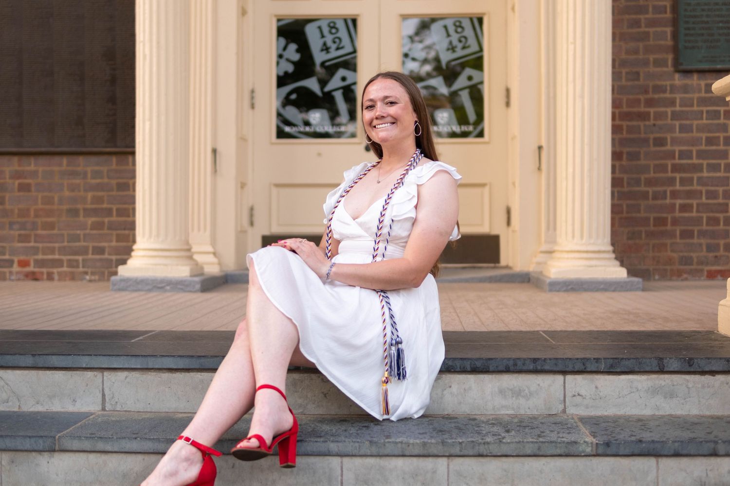 Smiling young woman in white dress and red heels sitting on steps of brick building.