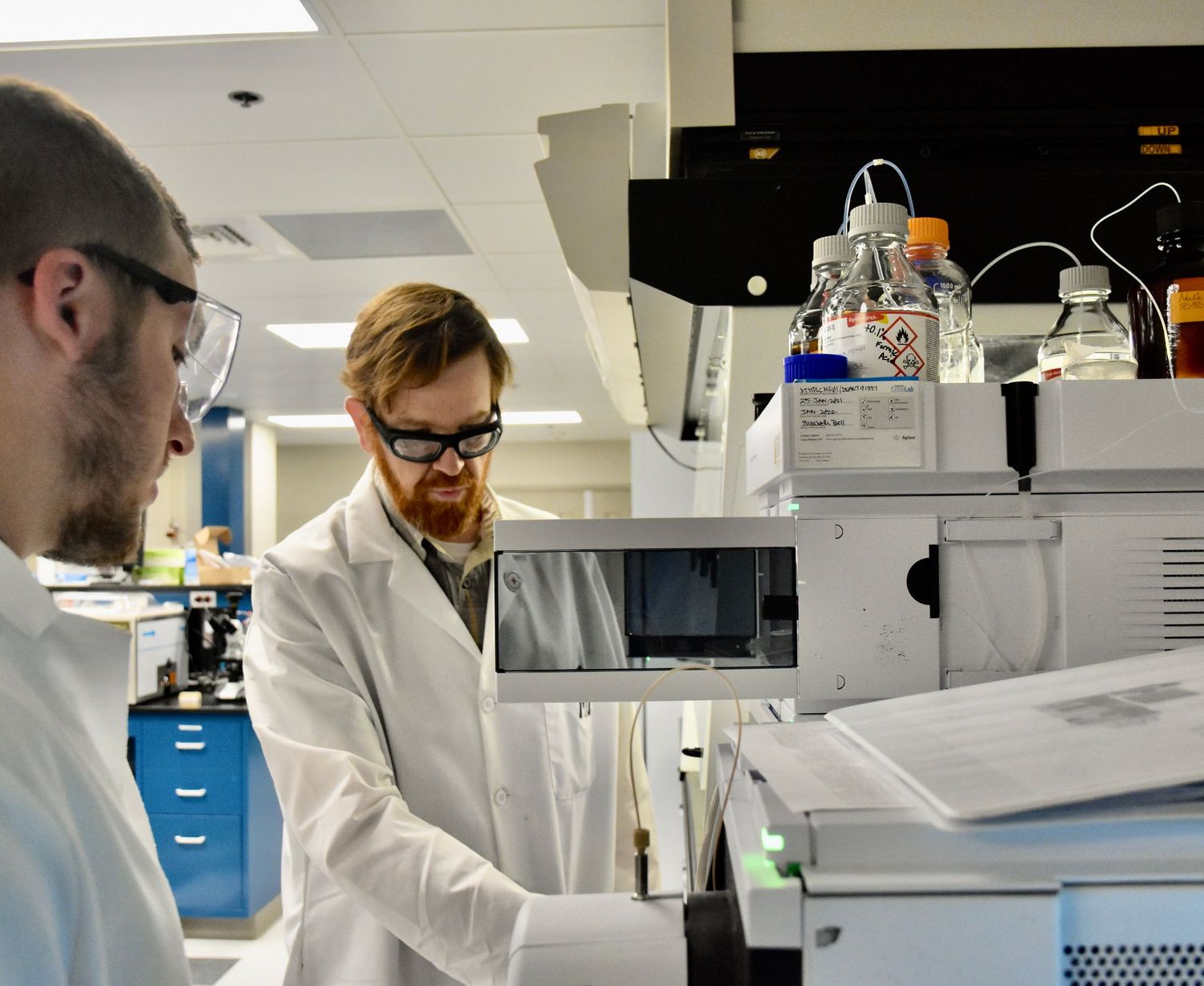 Two scientists in white lab coats and safety glasses work with laboratory equipment.