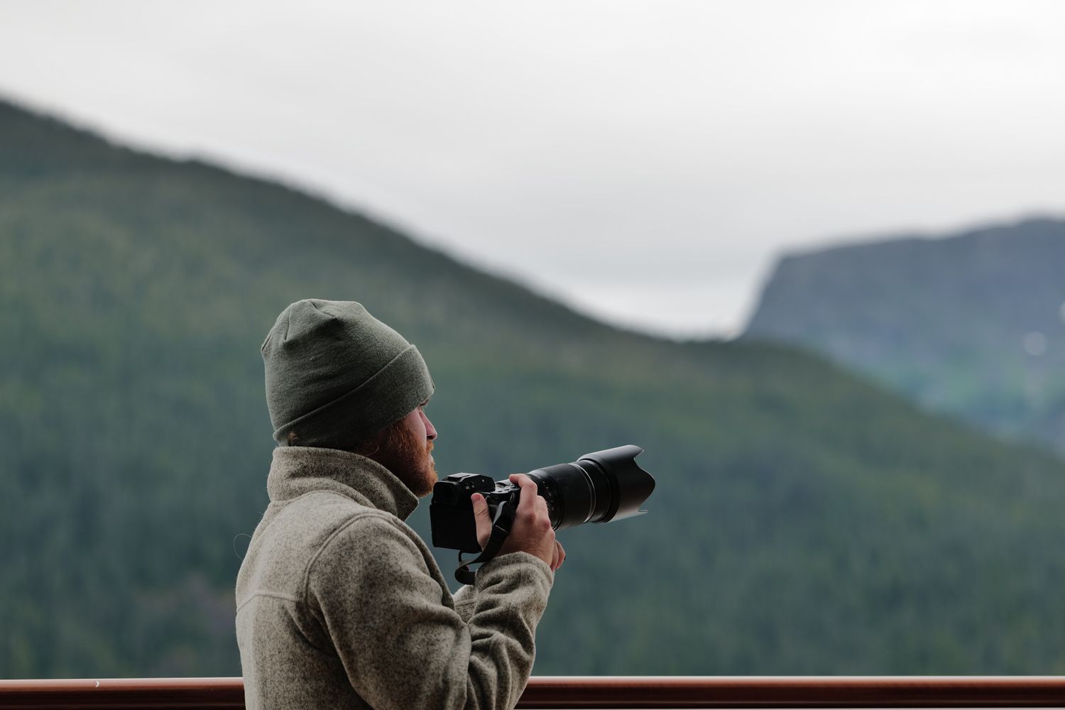 Photographer in a gray hoodie and beanie captures mountain landscape with DSLR camera on an overcast day.