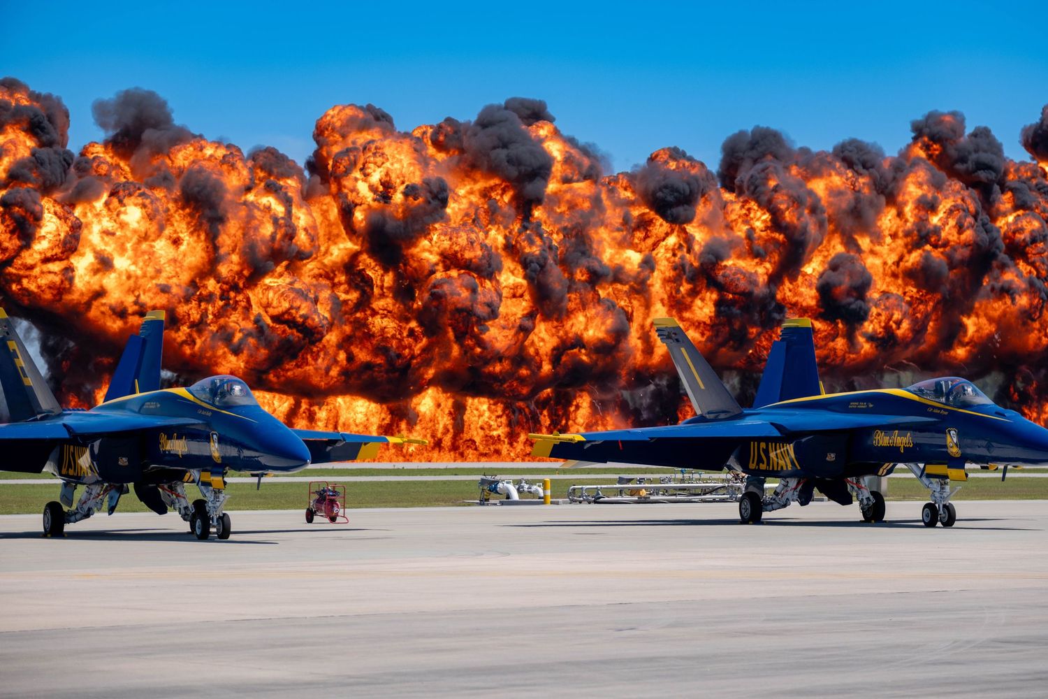 Two Blue Angels jets on the tarmac with a massive wall of fire and explosions behind them.