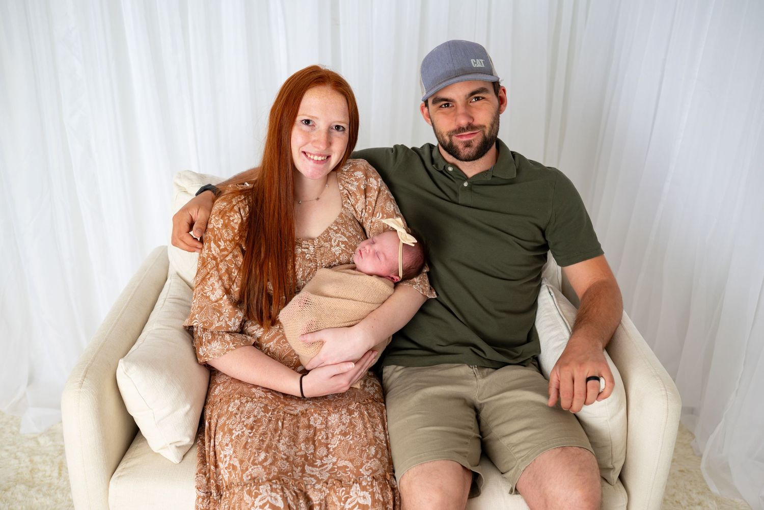 Happy couple sitting on white sofa holding their newborn baby, mother with long red hair wearing floral dress.