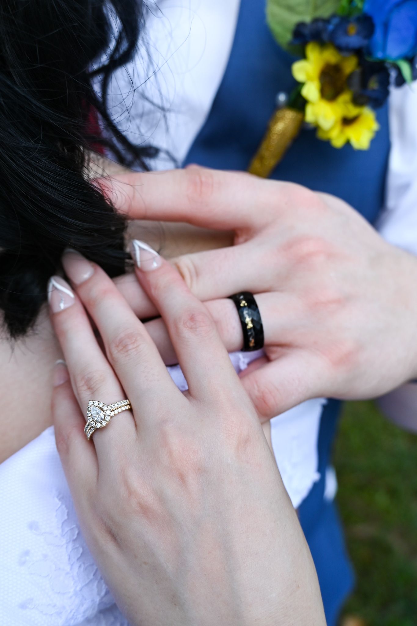 Newlywed couple displaying wedding rings, bride wearing diamond ring and groom wearing black band, with sunflower boutonniere.
