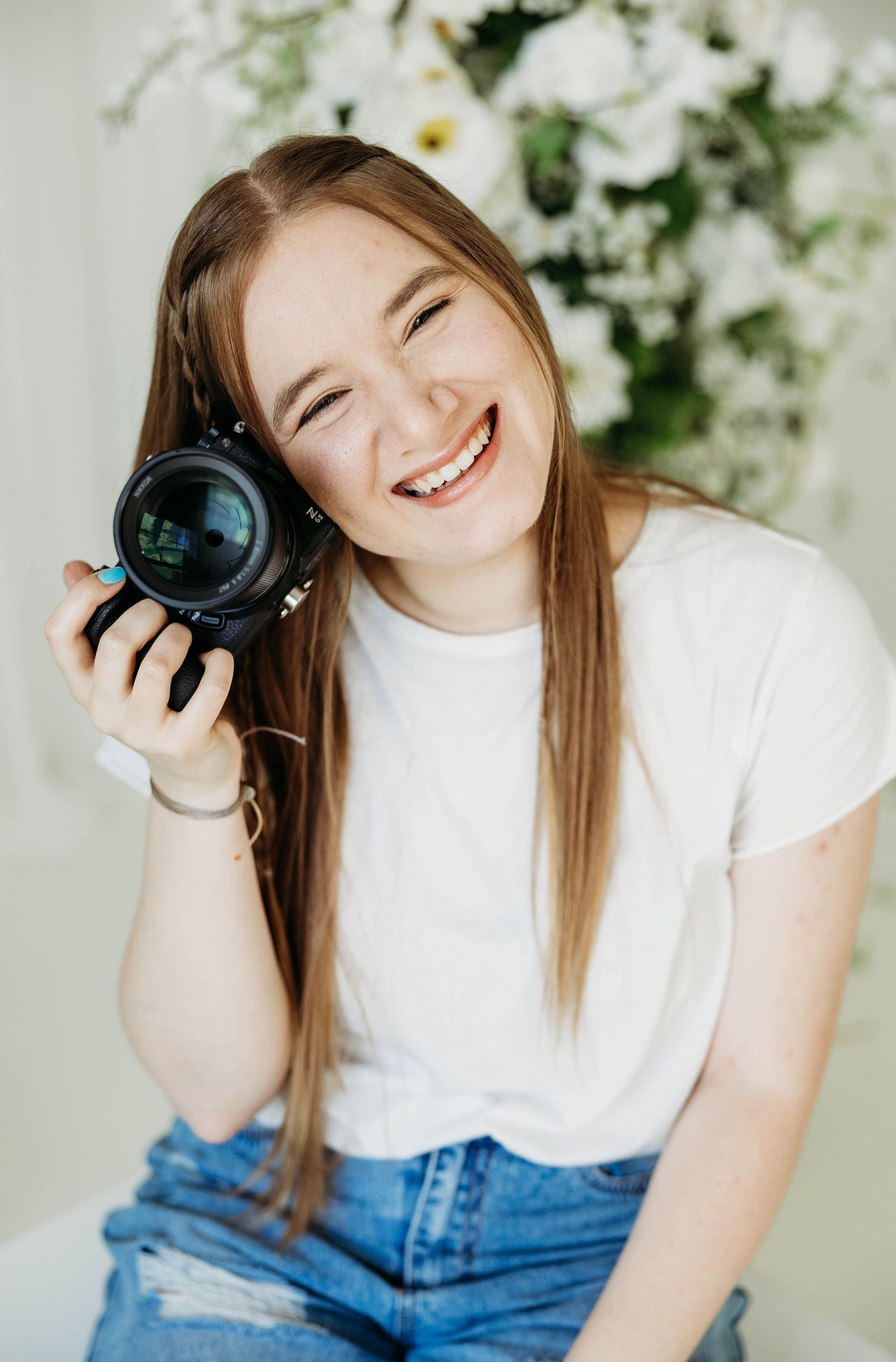 Smiling female photographer holding a camera beside white flowers in a bright studio.