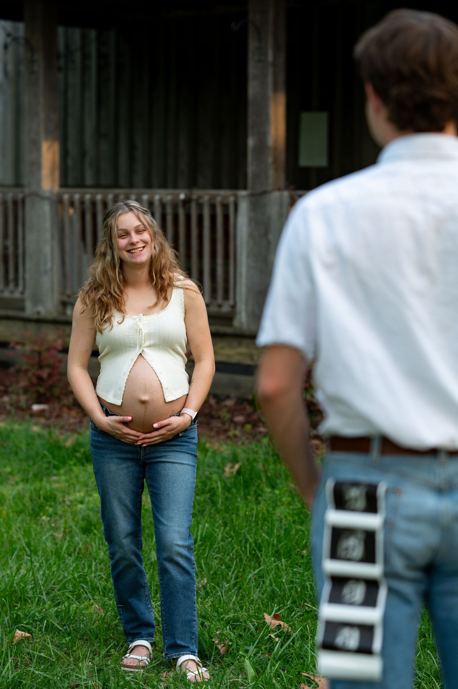 Smiling pregnant woman cradling her baby bump outdoors while her partner stands nearby holding ultrasound photos.