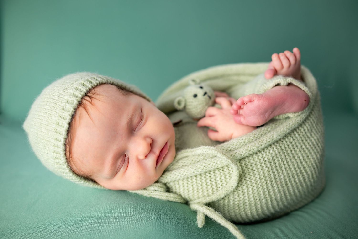 Sleeping newborn baby in white knit hat resting in green knitted basket with stuffed animal toy.