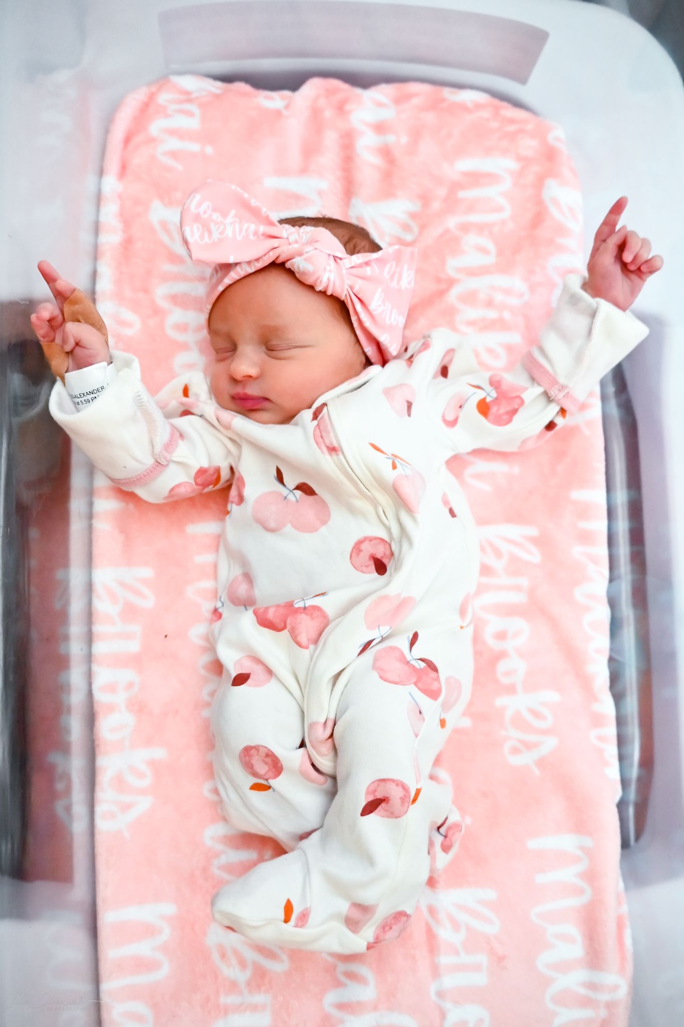 Sleeping newborn baby girl in peach floral outfit and pink bow headband lies on a personalized pink blanket at hospital.