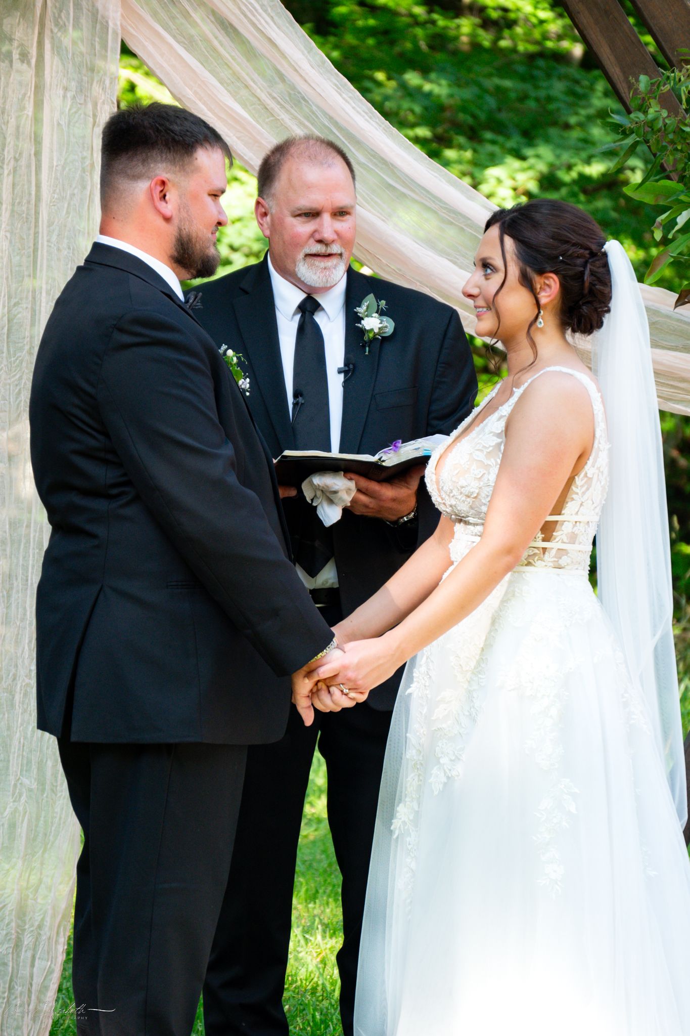 Bride and groom hold hands during outdoor wedding ceremony while officiant reads from book beneath draped canopy.