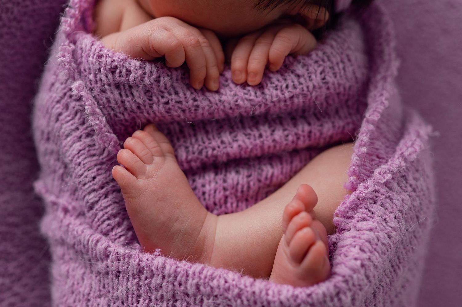 Newborn baby swaddled in a soft purple knit blanket, with tiny feet and hands visible.