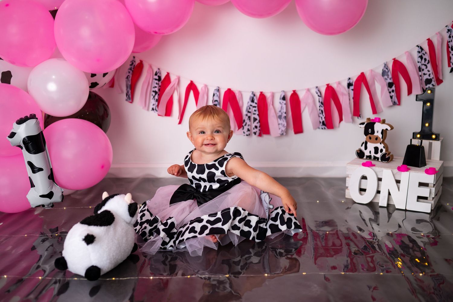 Baby girl in cow print tutu dress sits with pink balloons at first birthday party.