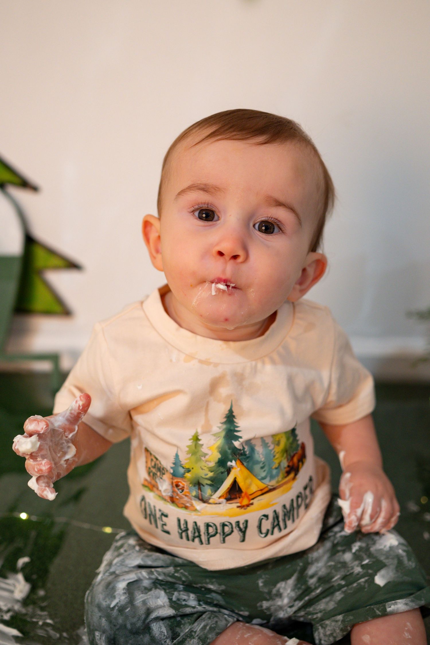 One-year-old in One Happy Camper shirt enjoys messy cake smash birthday photoshoot.