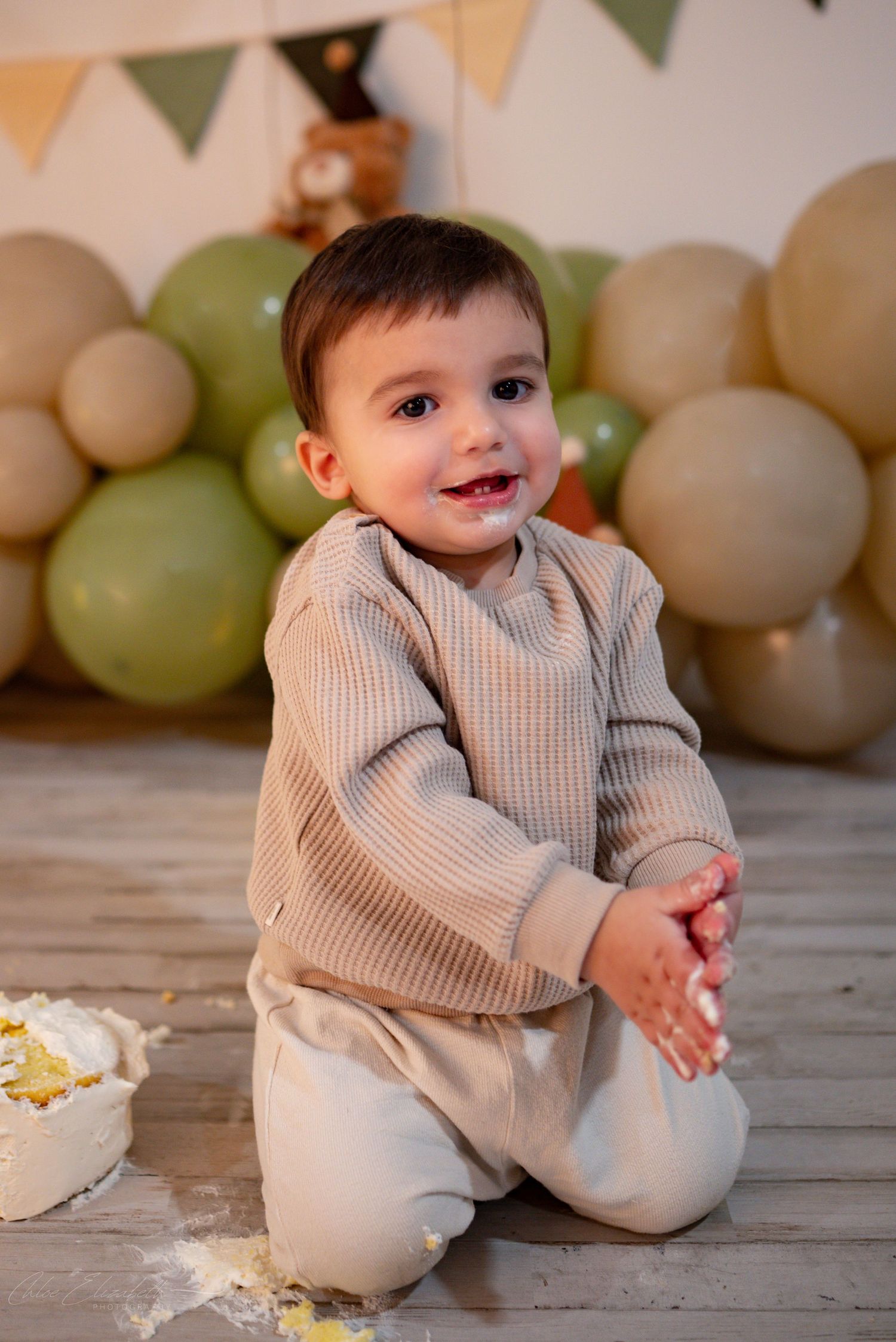 Smiling one-year-old boy with cake on hands kneeling by smashed birthday cake and balloons.