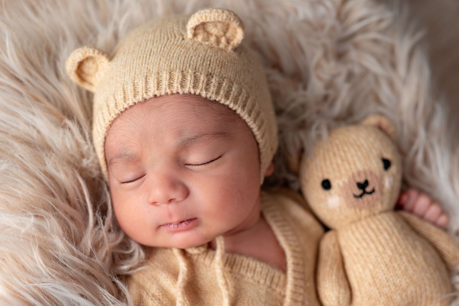 Close-up of sleeping newborn wearing beige bear ear hat snuggled next to knitted teddy bear.
