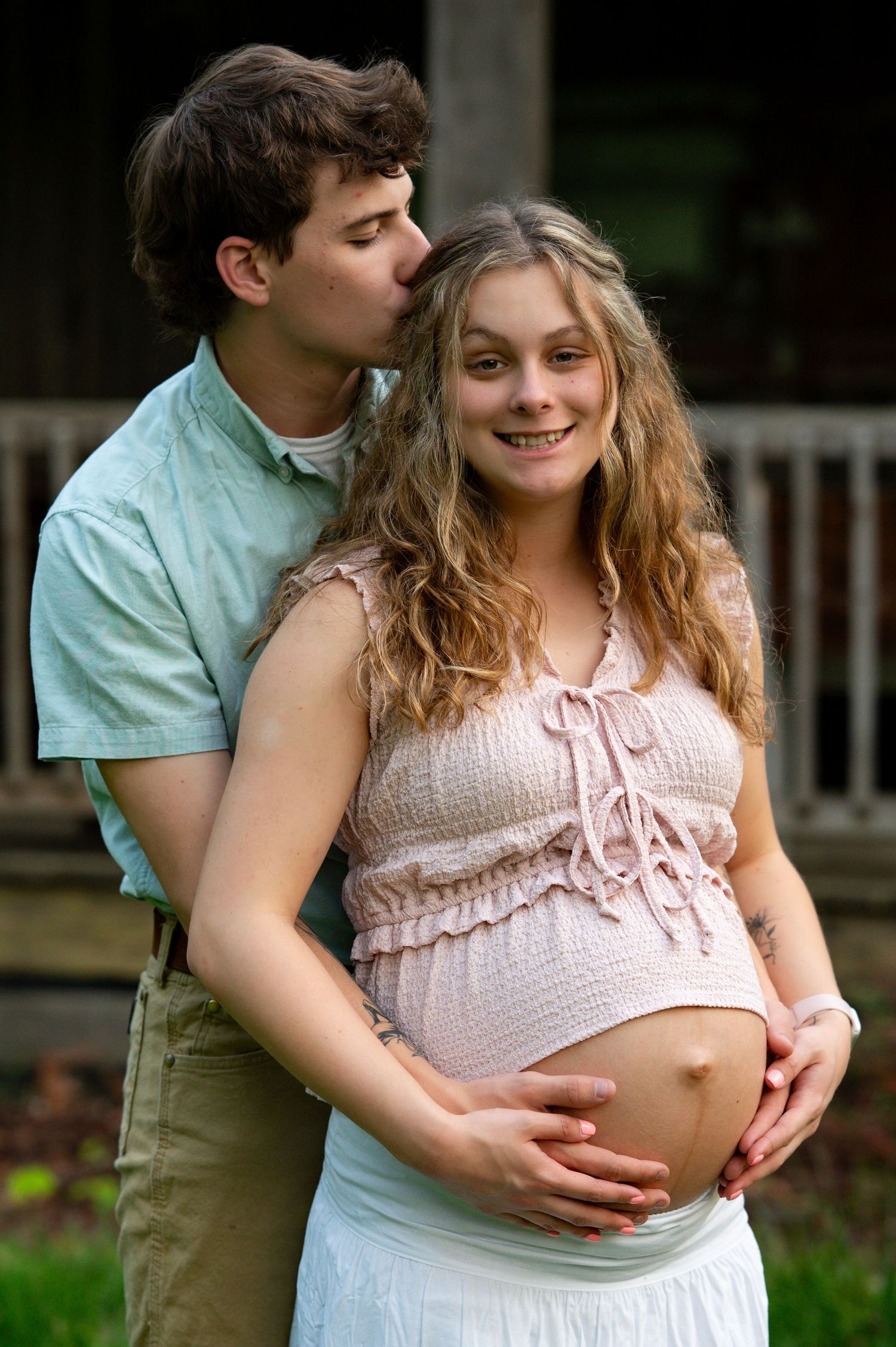 Man kissing pregnant woman on the cheek from behind while both cradle her bare baby bump outdoors.