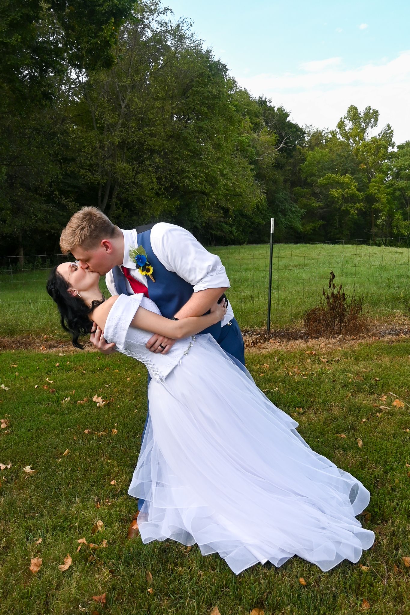 Bride and groom share a romantic dip kiss outdoors on green grass with trees in background at wedding.