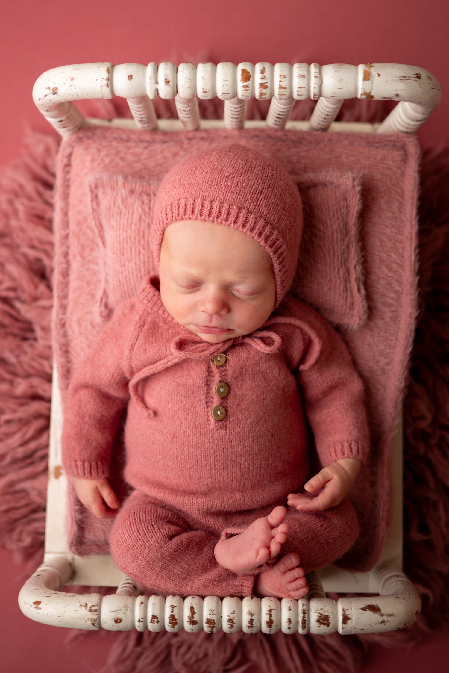 Newborn baby sleeping in pink knit outfit and bonnet on a small white vintage bed with pink fur blanket.