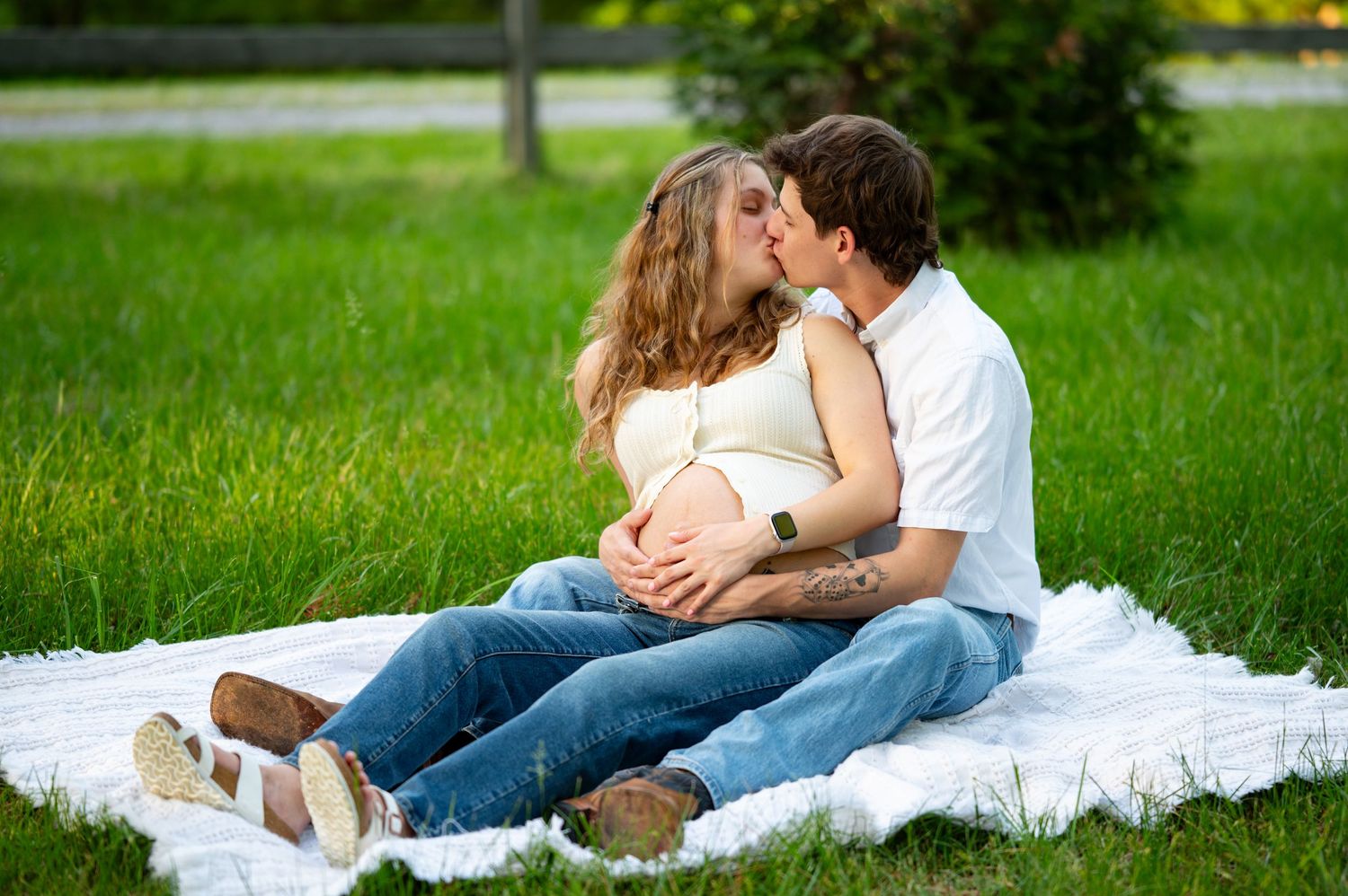 Pregnant couple kissing on a blanket in a green park, hands on baby bump.