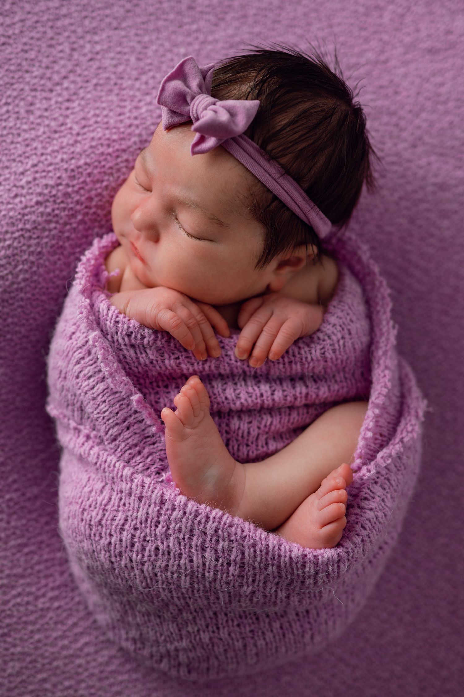 Newborn baby girl swaddled in purple knit wrap with bow headband, sleeping peacefully on lavender background.