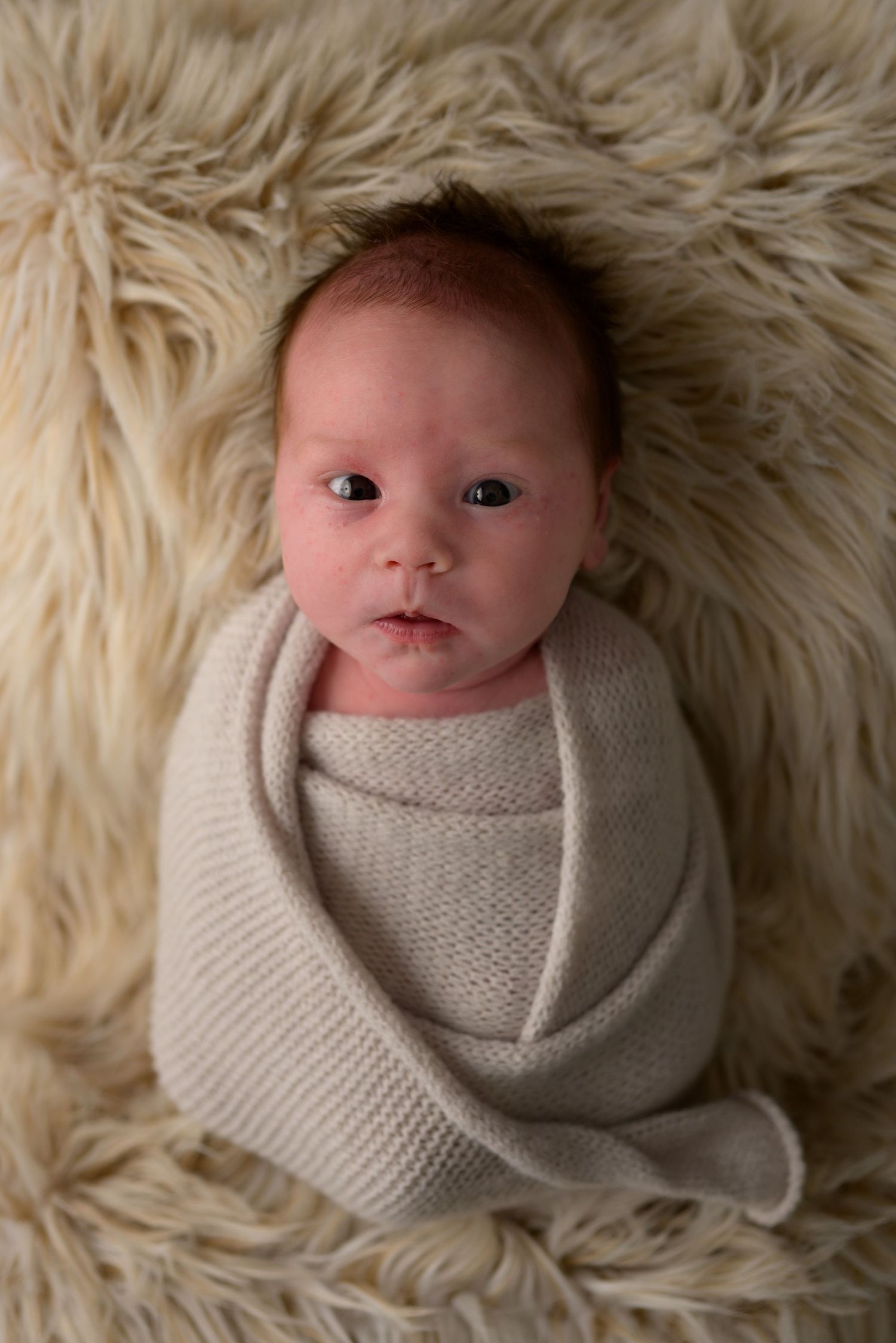 Newborn baby swaddled in a soft beige knit wrap, lying on a fluffy cream fur blanket during a professional photo session.