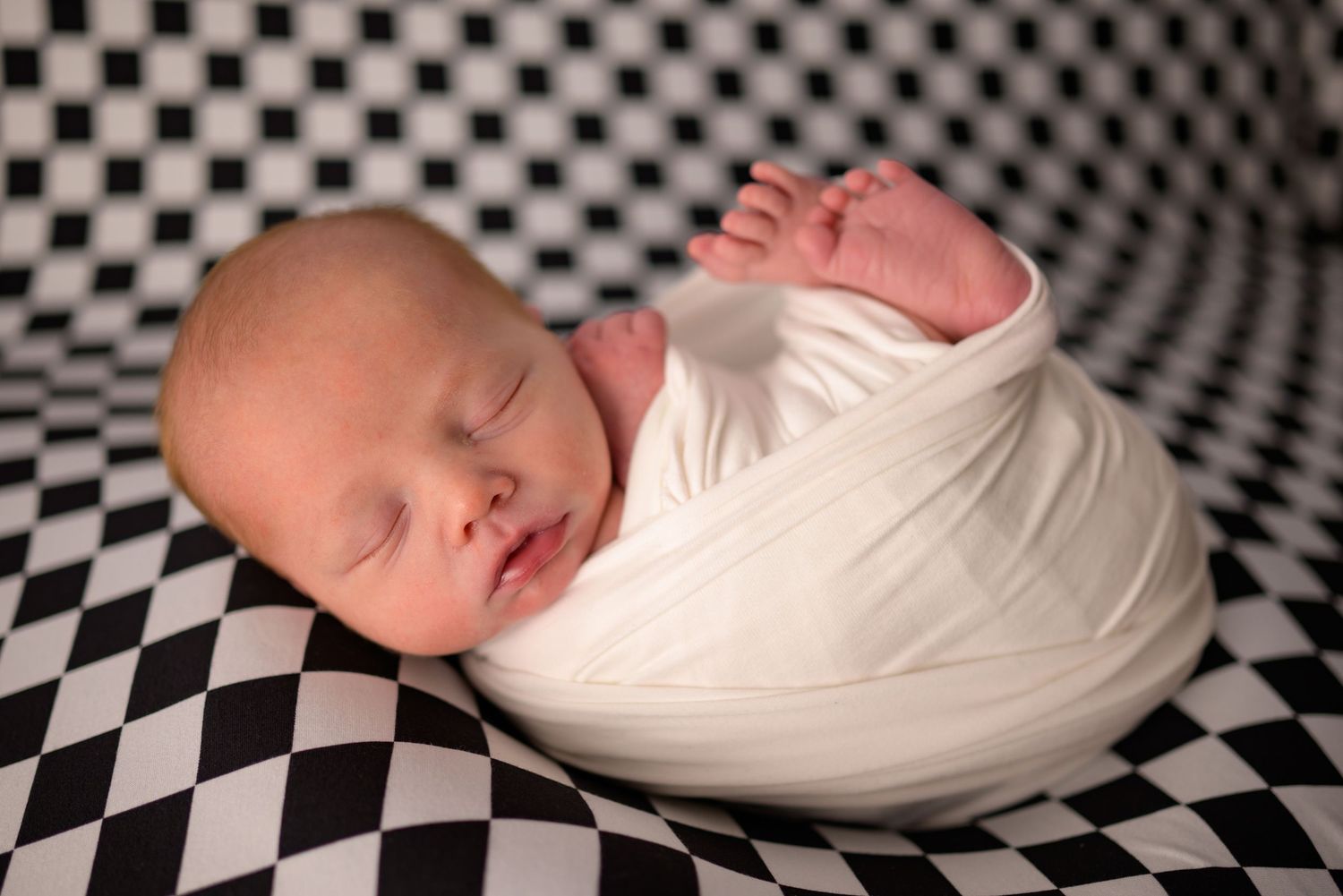 Newborn baby swaddled in white wrap sleeping peacefully on black and white checkered background.