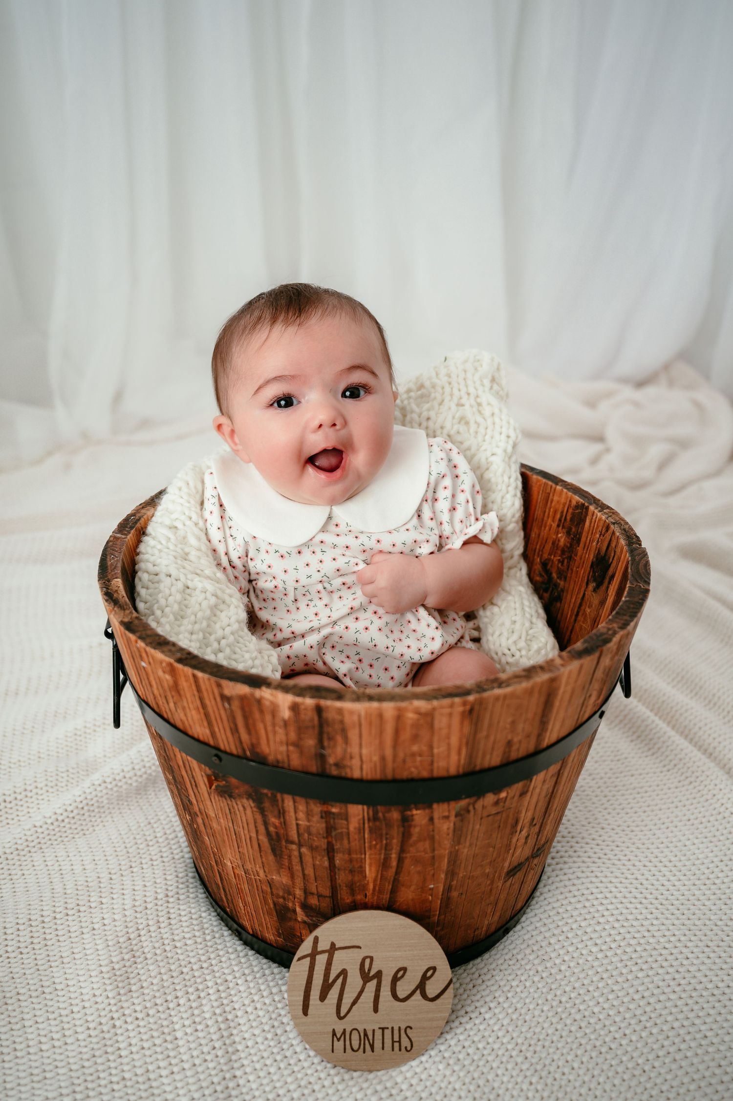 A happy 3-month-old baby sitting in a rustic wooden barrel bucket, wearing a white floral romper.