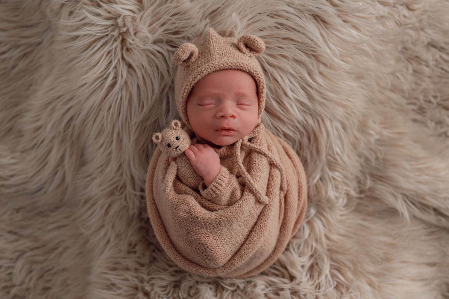 Newborn baby swaddled in beige knit wrap and bear hat, sleeping peacefully on fluffy fur blanket holding tiny teddy bear.