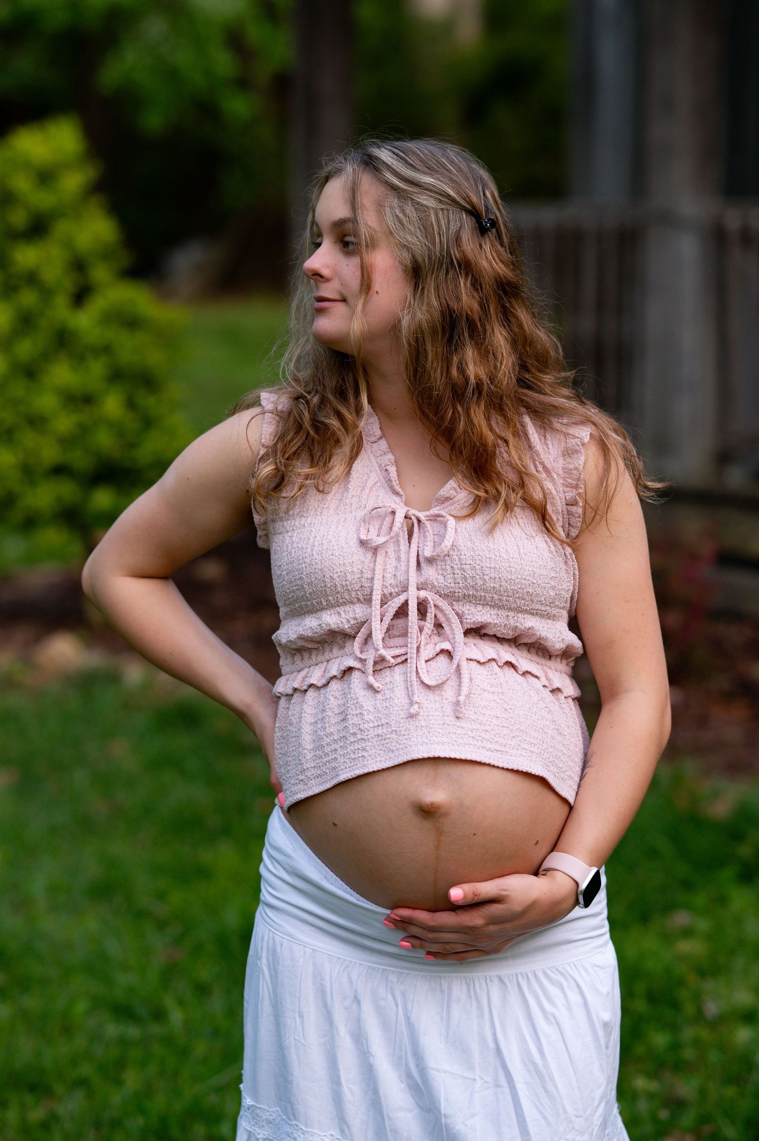 Pregnant woman in pink crop top and white skirt cradling her baby bump while looking away outdoors.