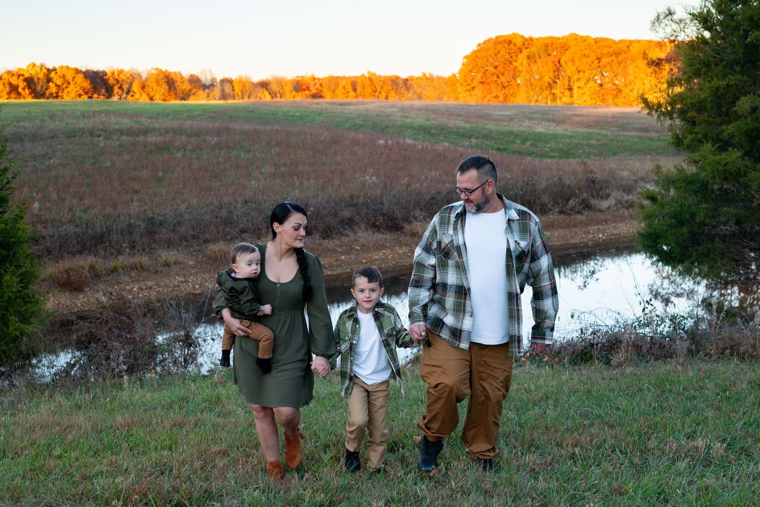 Family of four walking in golden autumn field near pond at sunset, children in coordinating outfits.