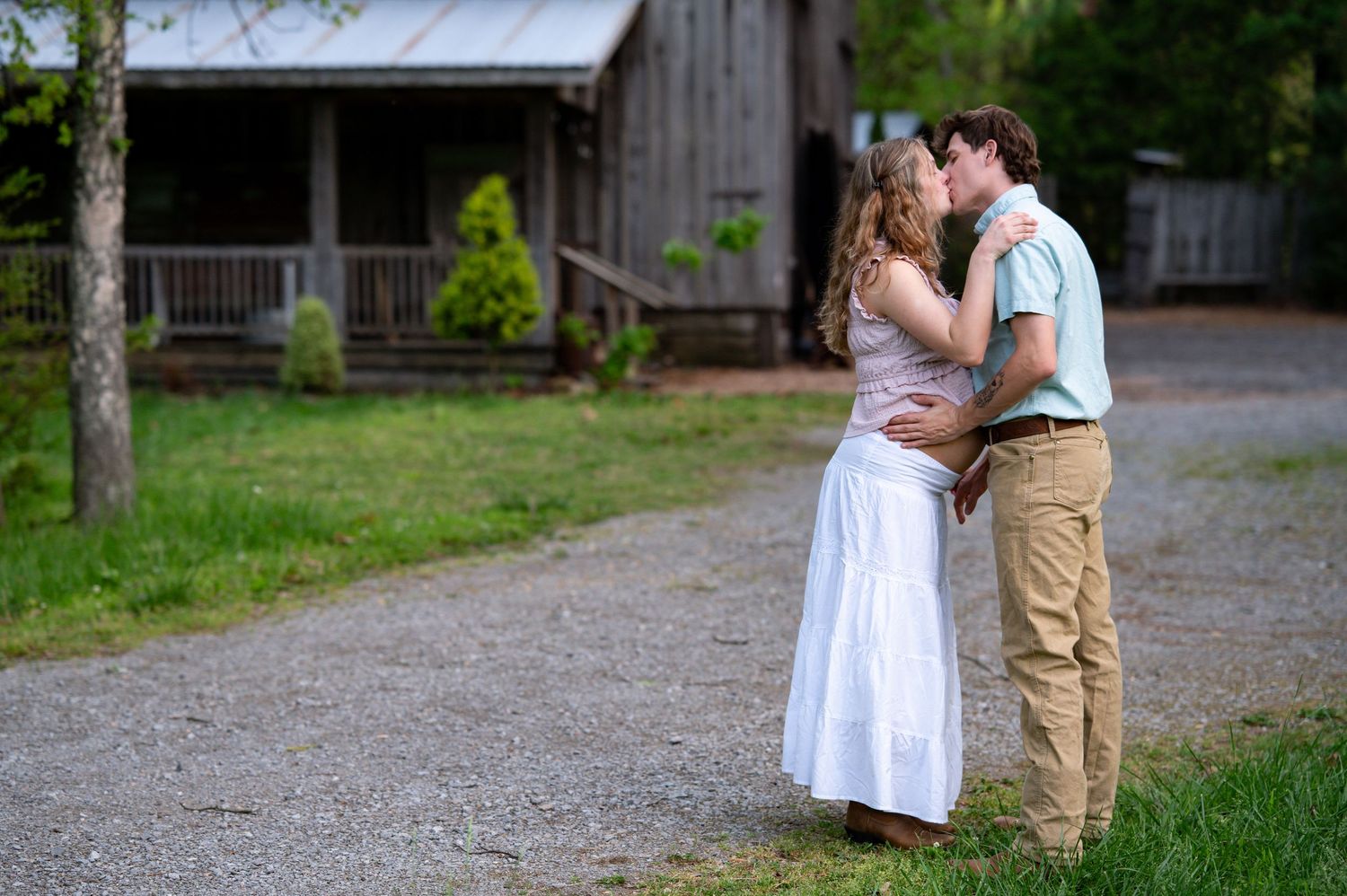 Couple sharing a romantic kiss on a gravel path near a rustic barn, woman in white dress.