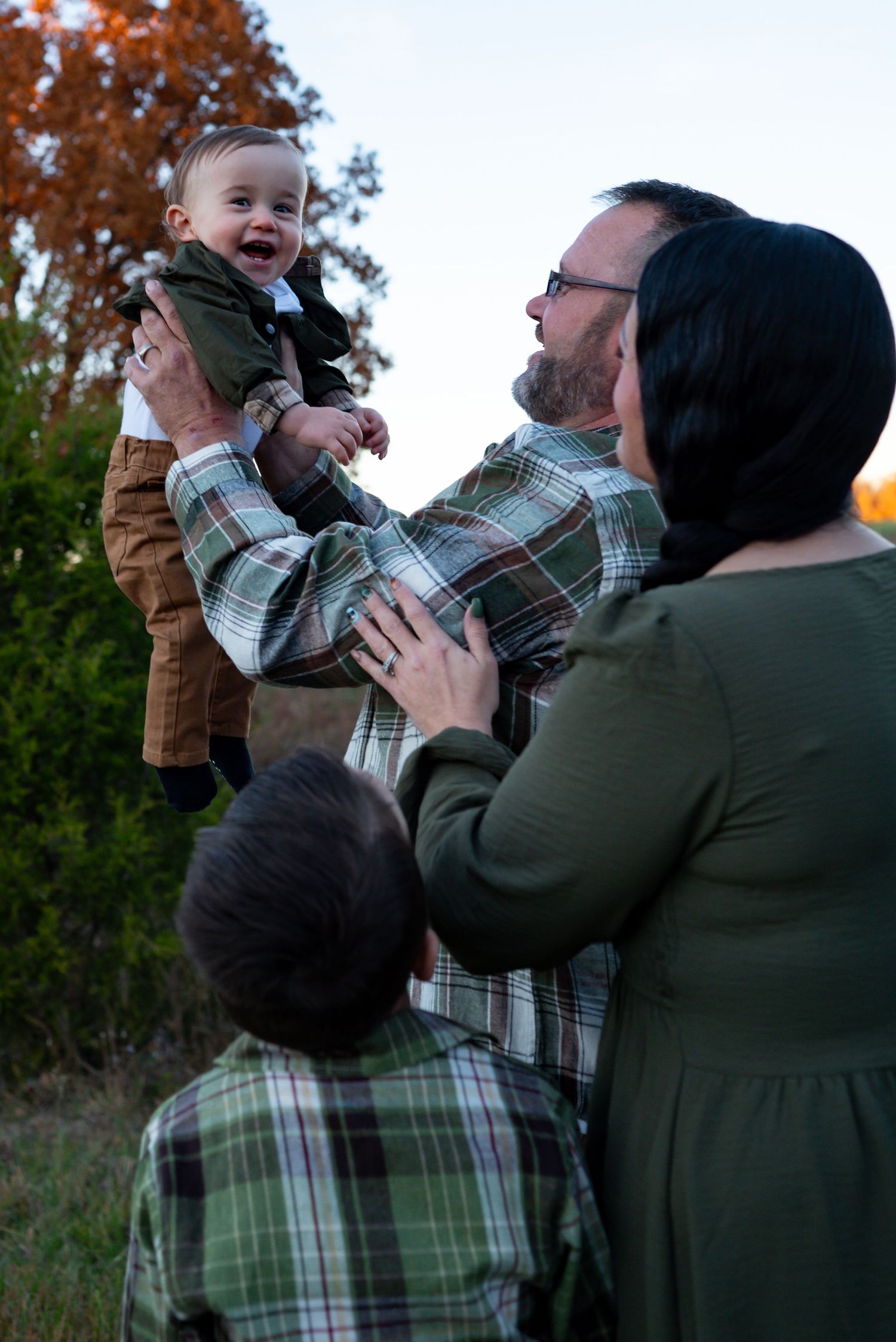 Family outdoor portrait with father lifting laughing baby while mother and young boy look on among autumn foliage.