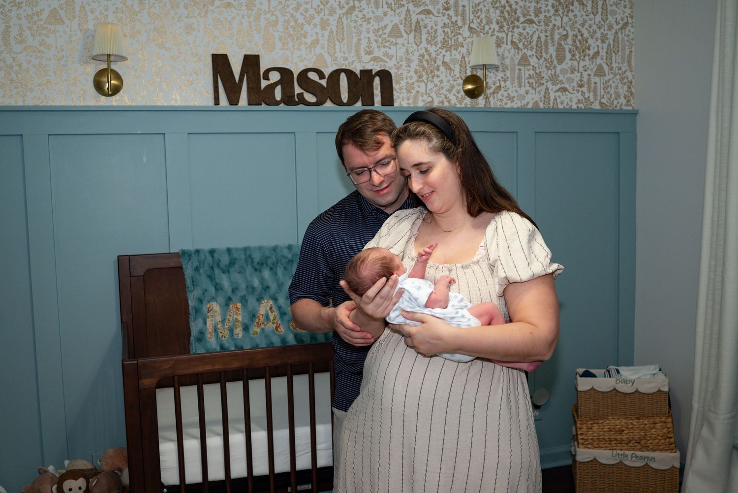 Parents admire newborn baby in nursery with blue walls and wooden Mason name sign above crib.