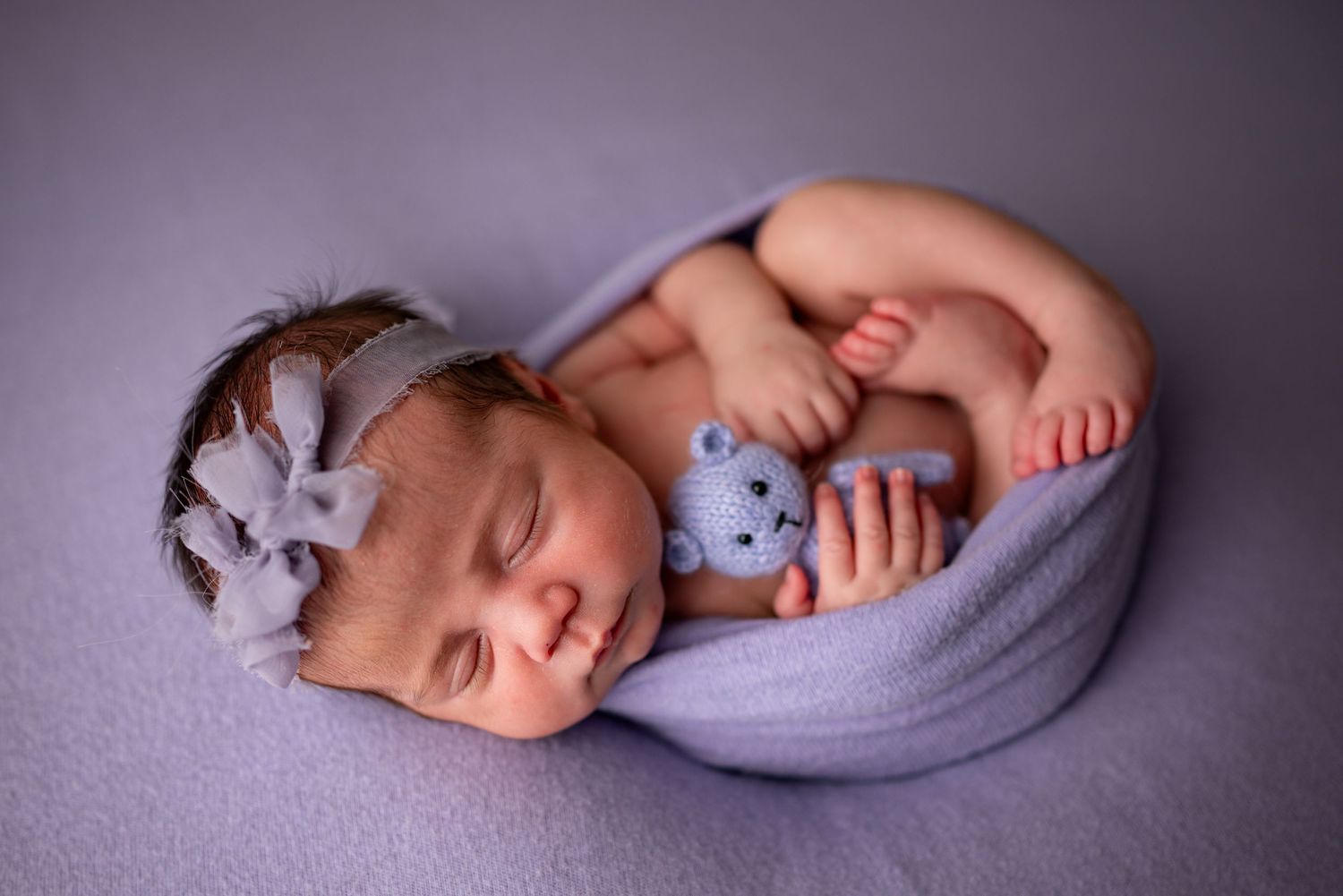 Newborn baby girl sleeping peacefully wrapped in lavender, holding a tiny knit bear, wearing a purple bow headband.