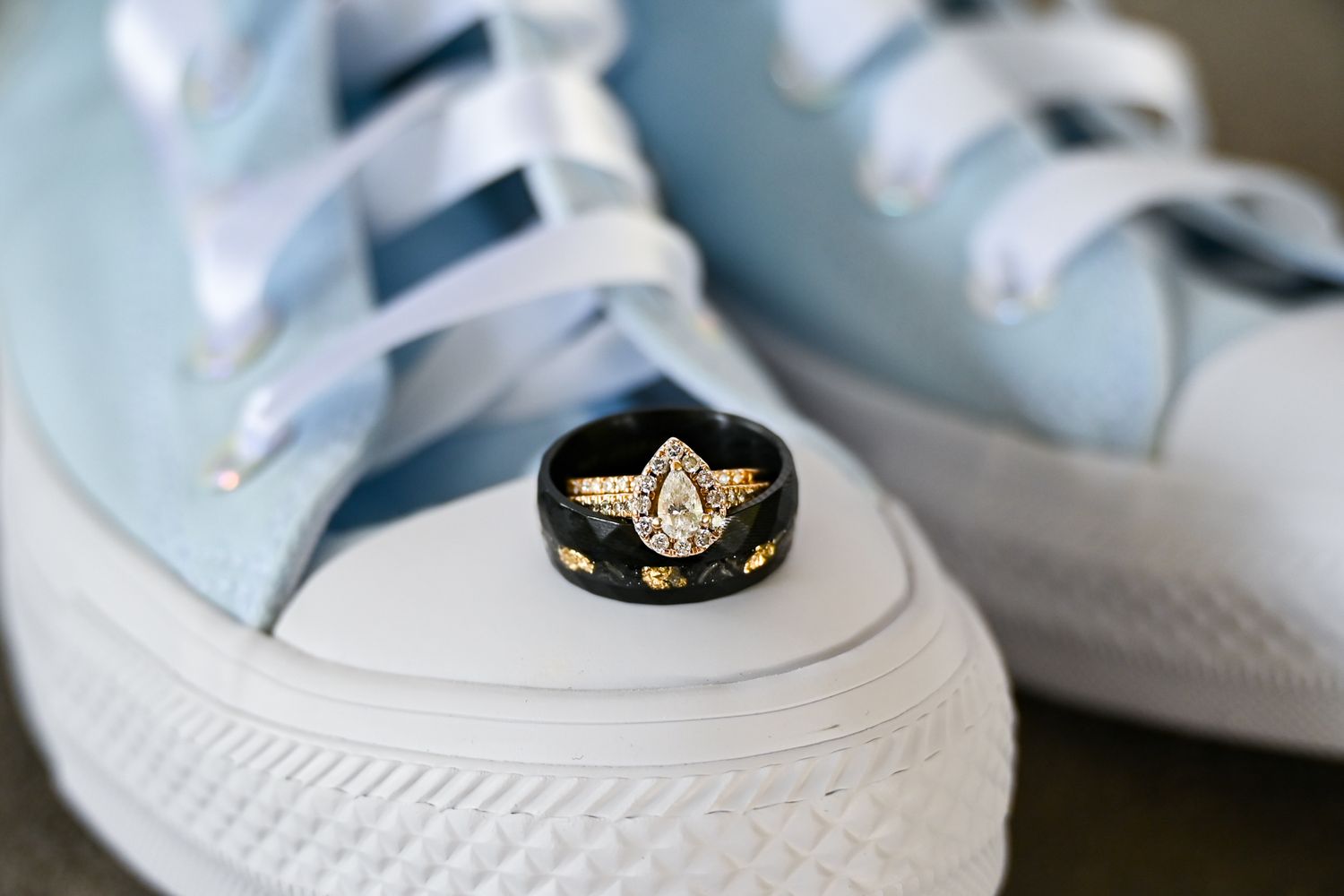 Gold pear-shaped diamond engagement ring with black band displayed on light blue Converse sneakers.