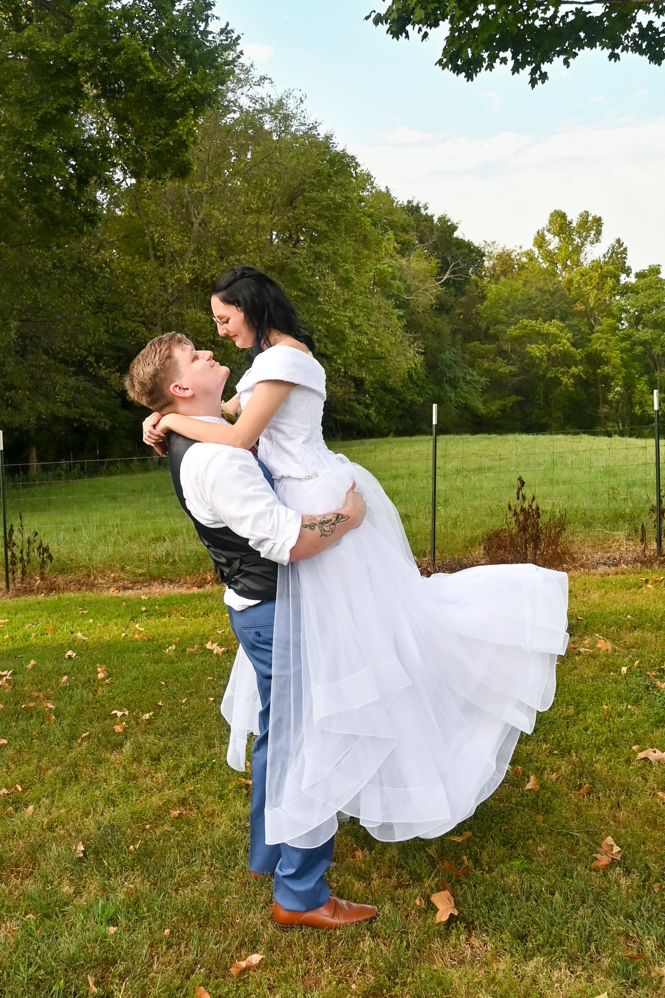 Groom lifts bride in white wedding dress outdoors in lush green park setting, sharing a romantic moment.