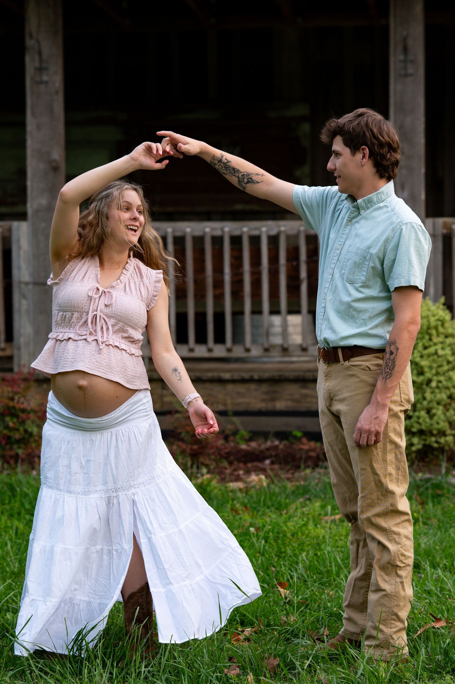 Expecting couple dancing and laughing together on green grass in front of a rustic wooden porch.