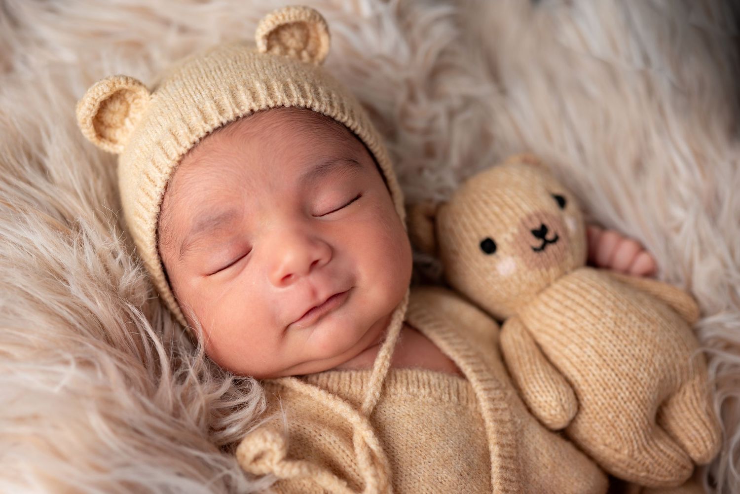 Newborn baby sleeping peacefully in tan bear ear bonnet cuddled next to matching knit teddy bear.