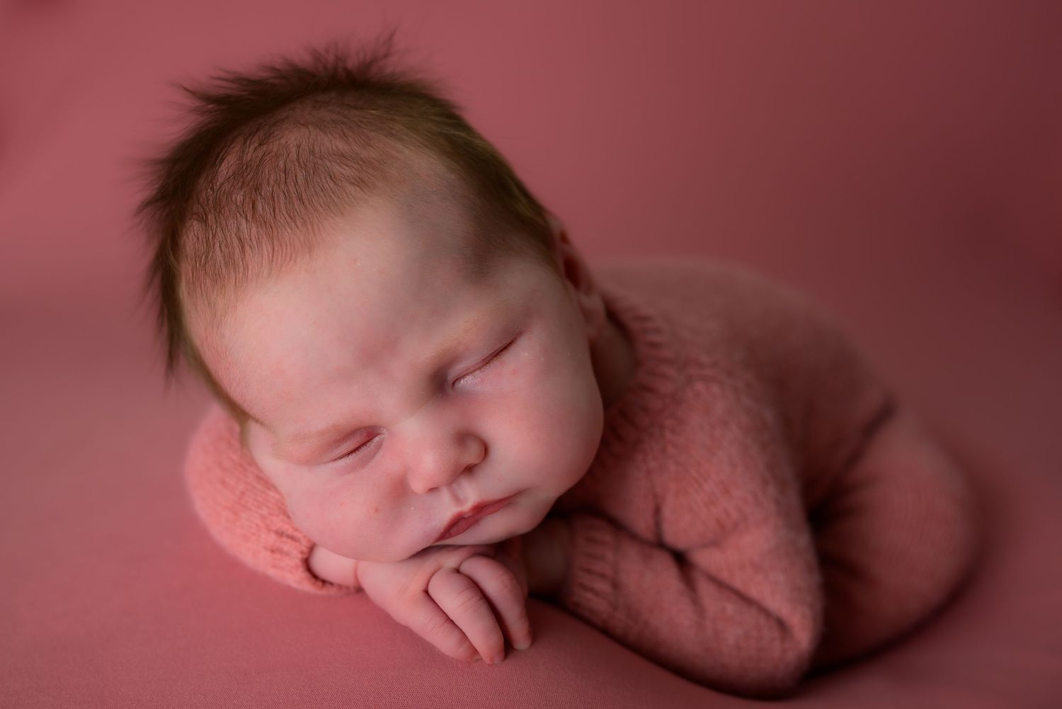 Sleeping newborn baby in pink knit outfit posed on pink backdrop in professional newborn photography session.