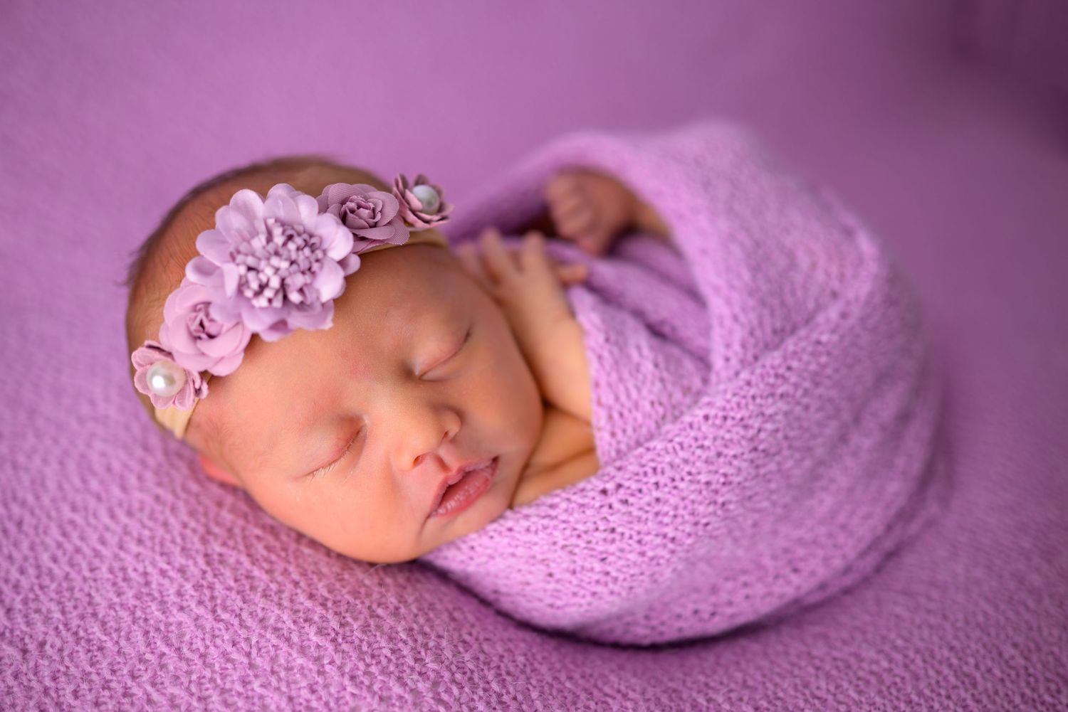 Peaceful sleeping newborn girl wrapped in purple knit swaddle wearing a delicate floral headband.