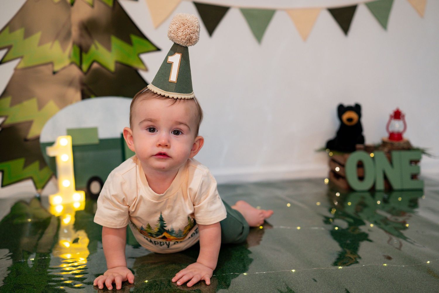 Baby boy in camping hat celebrates first birthday with woodland-themed cake smash decorations.