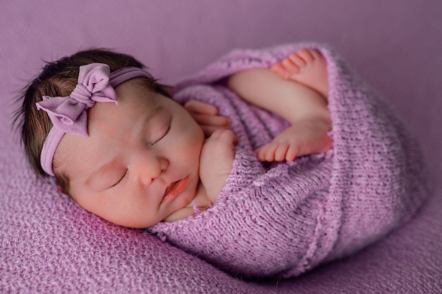 Newborn baby girl sleeping peacefully wrapped in purple knit blanket with matching bow headband.