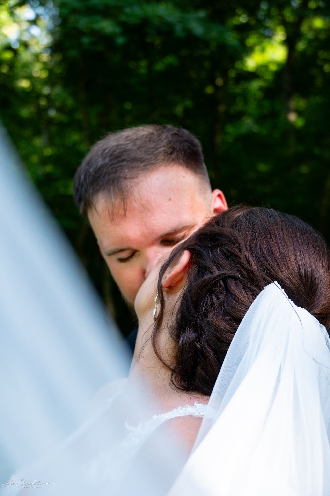 Groom tenderly kissing bride on her head during outdoor wedding ceremony, surrounded by lush green trees.