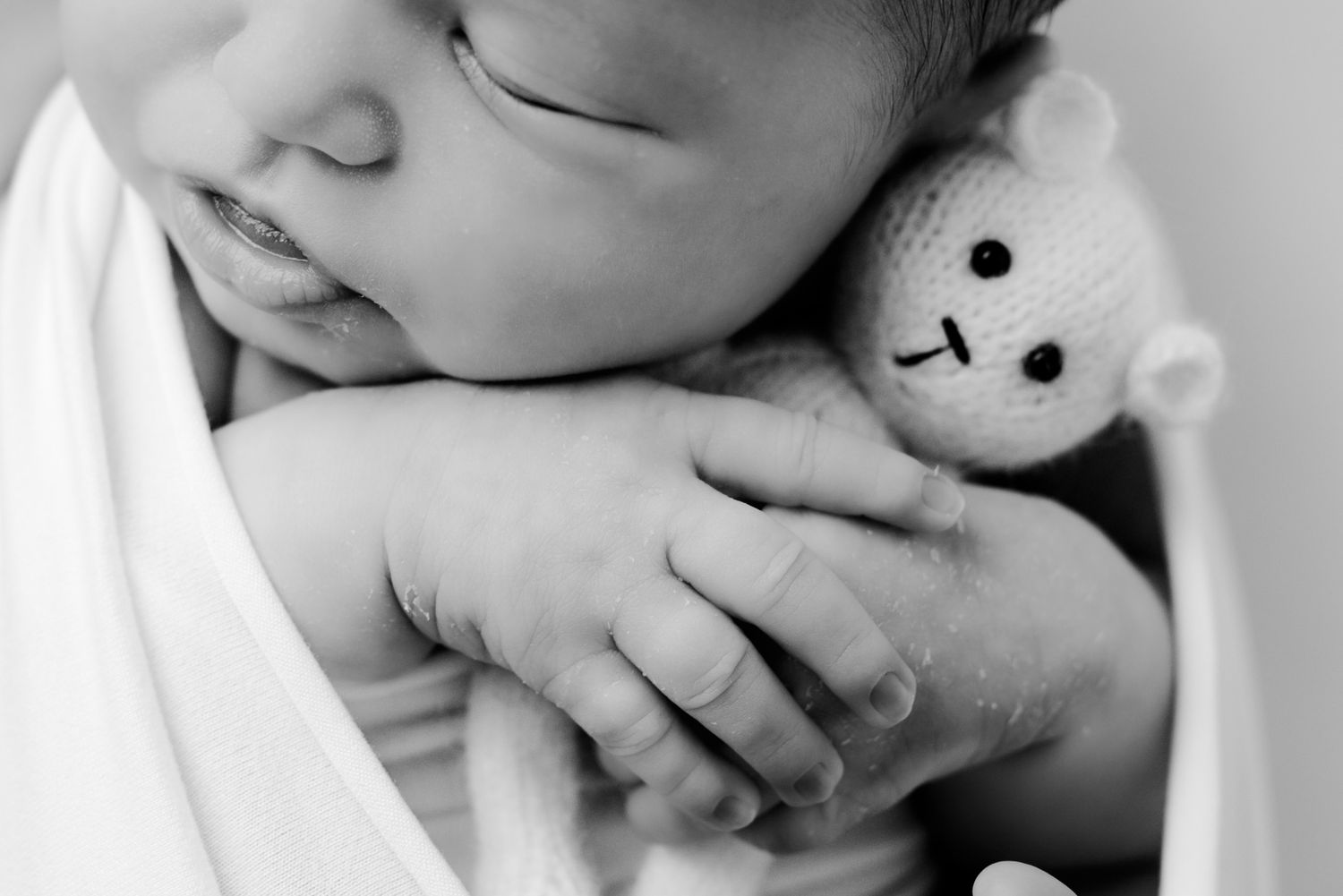 Close-up black and white photo of sleeping newborn baby holding a tiny knit teddy bear.