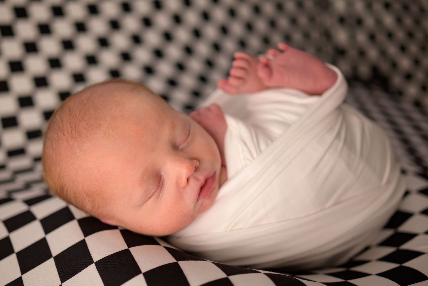 Peacefully sleeping newborn wrapped in white swaddle on black and white checkered surface.