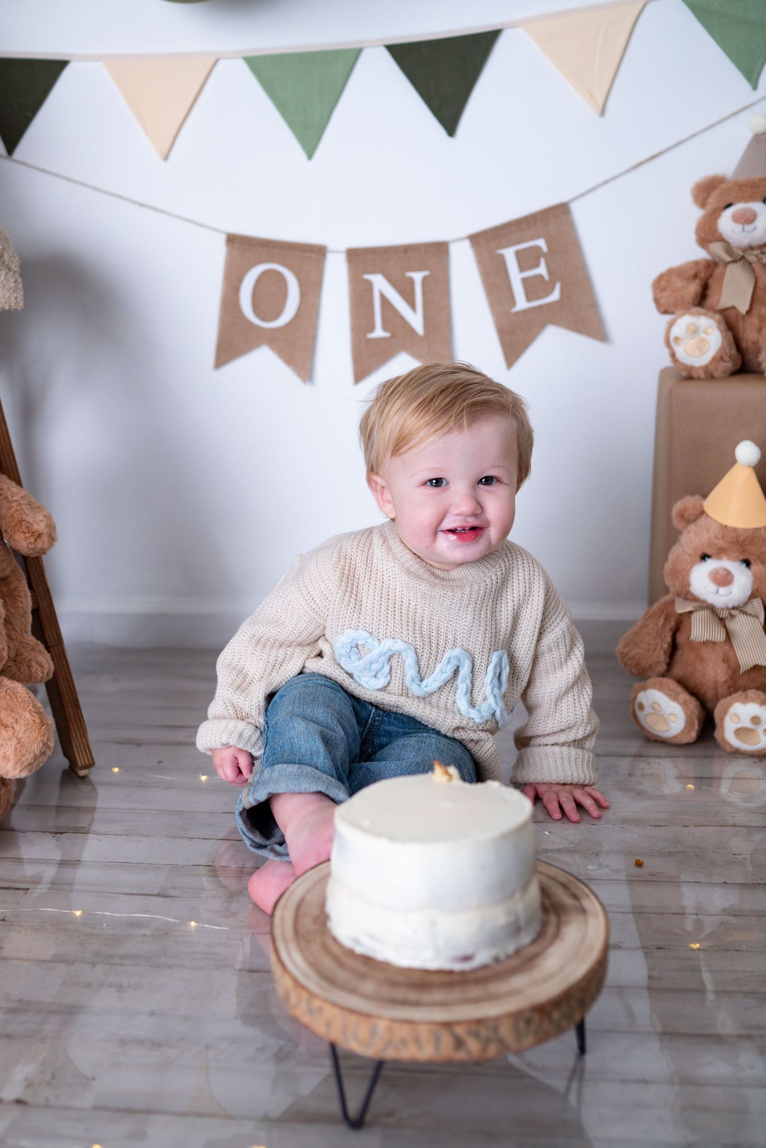 Happy one-year-old boy smiles at camera beside white birthday cake with ONE sweater and banner decor.