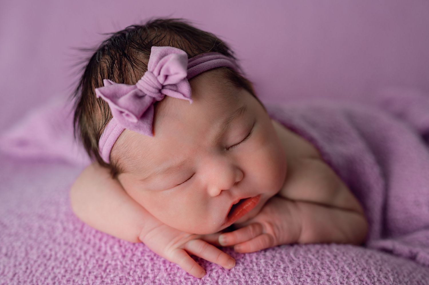 Newborn baby girl sleeping peacefully wearing a purple bow headband, wrapped in a soft lavender blanket.