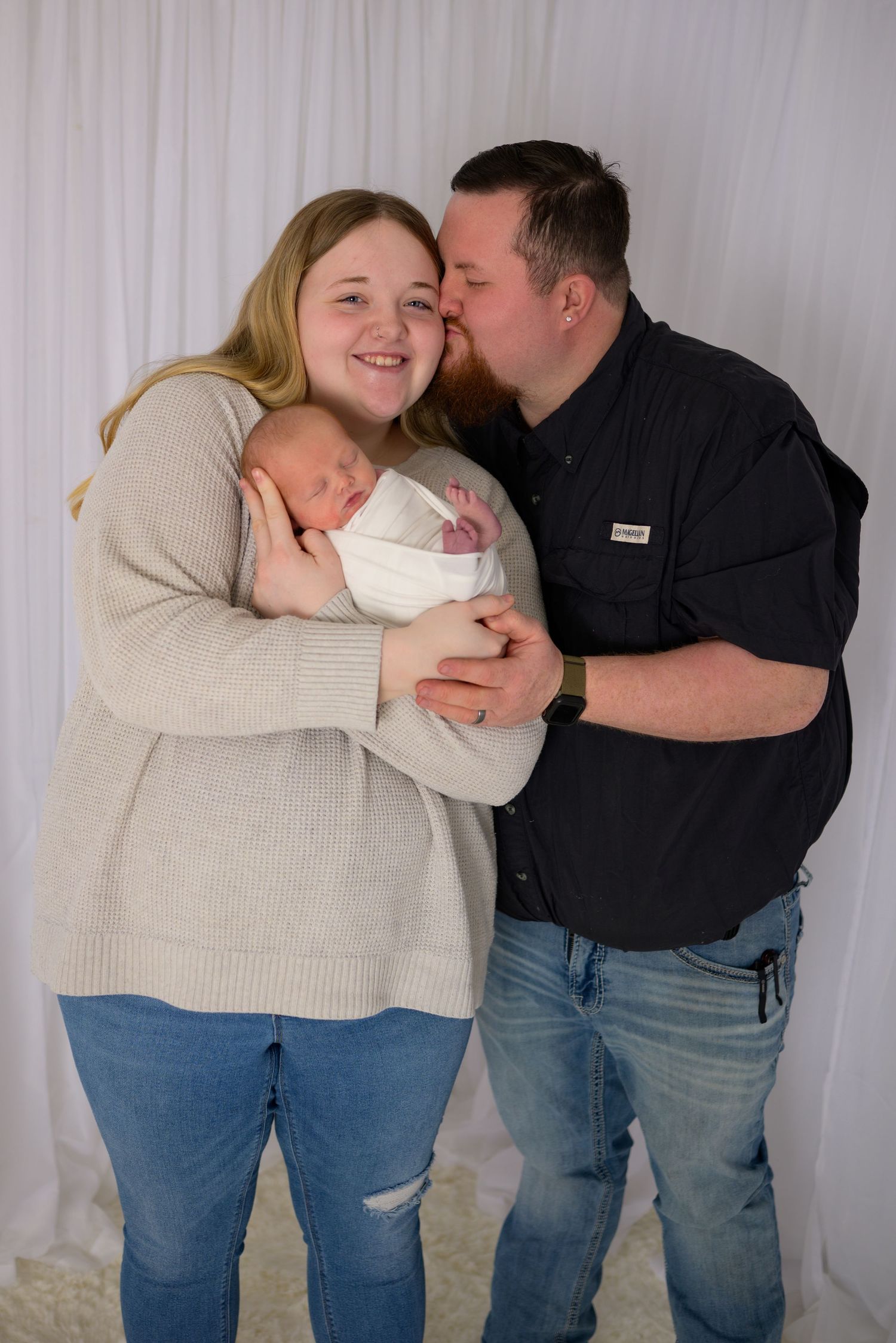 Couple smiling as father kisses mother on cheek while she holds swaddled newborn baby.