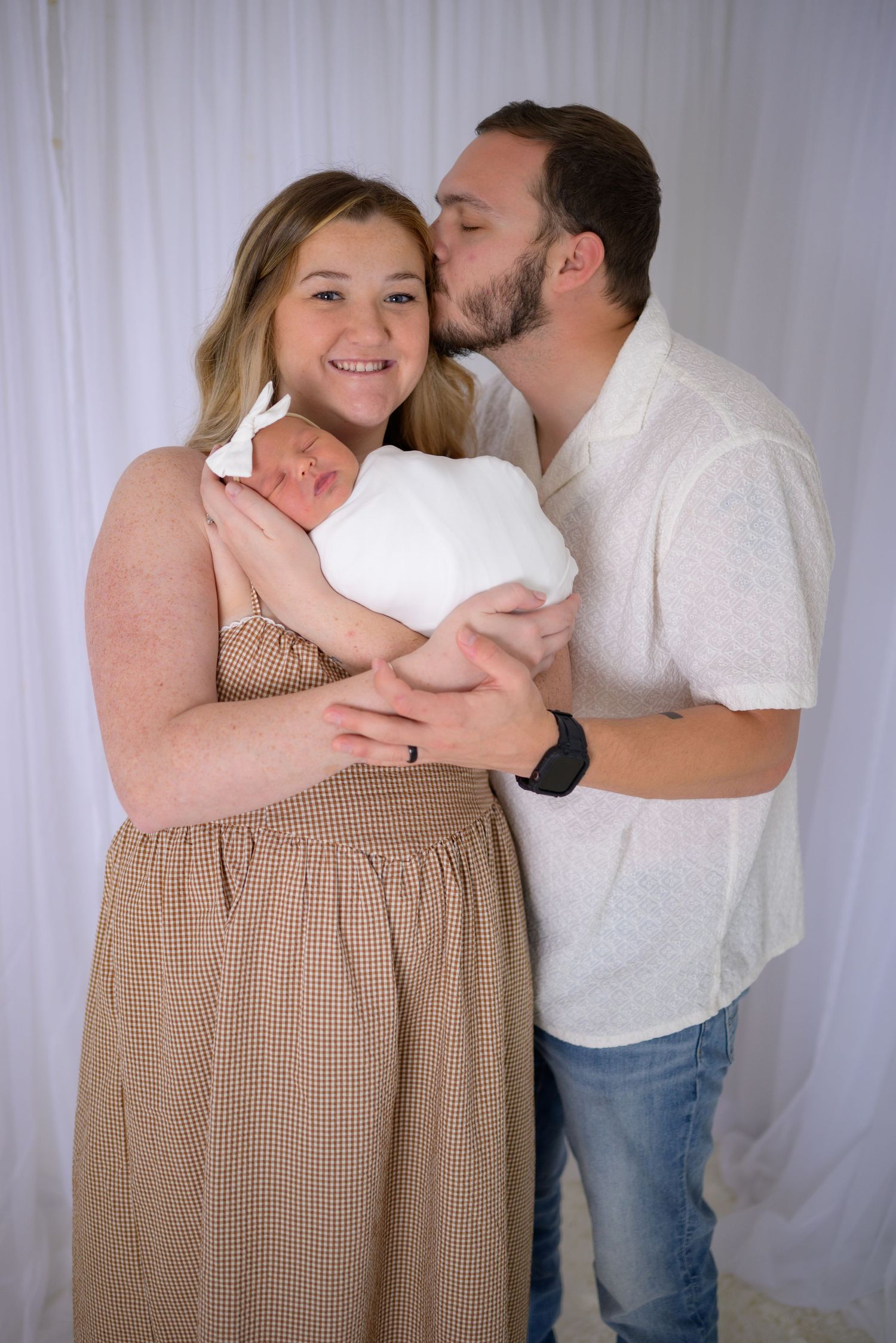 Young couple posing with swaddled newborn baby, man kissing womans cheek in white backdrop studio.