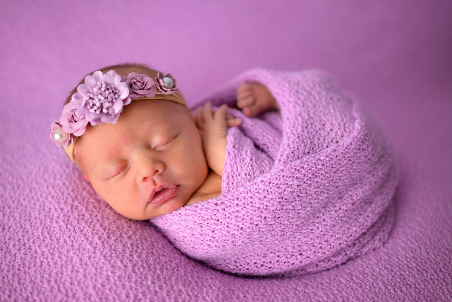 Newborn baby girl sleeping in purple knit wrap with decorative flower headband on purple background.