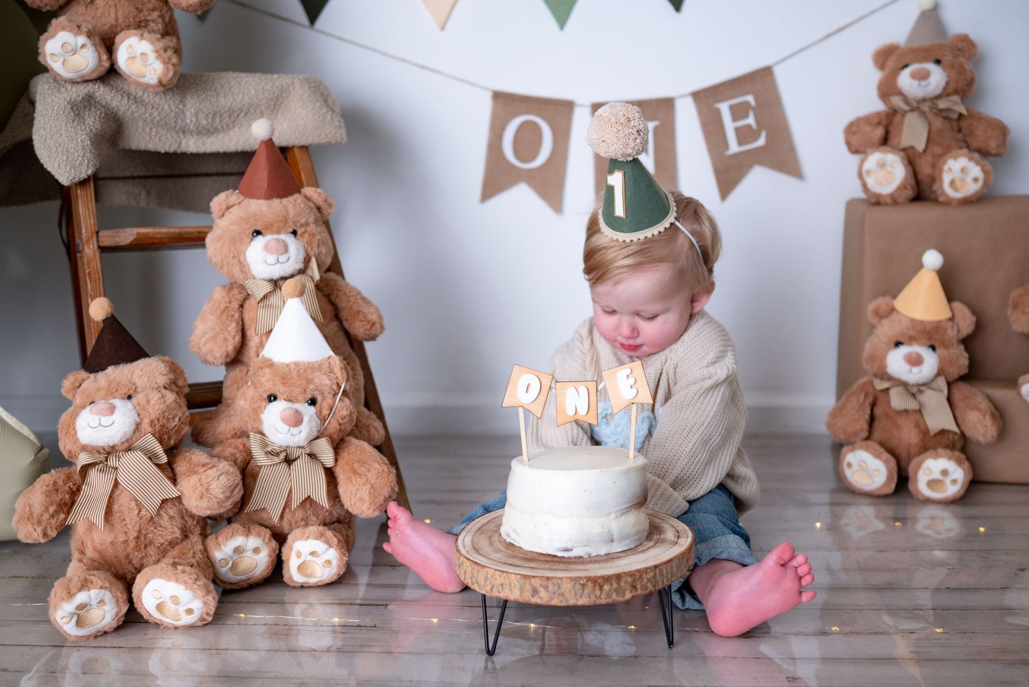 One-year-old boy leans toward smash cake surrounded by teddy bears and ONE banner backdrop.
